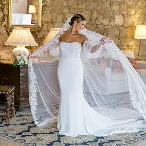 Noiva sorridente a segurar um véu longo de renda num salão rústico com paredes de pedra, fotografada em Portugal por Boris the Cat.