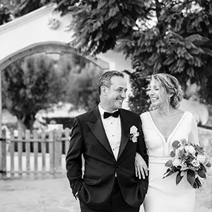 Casal de noivos a caminhar e sorrir de braço dado num retrato a preto e branco, estilo fotojornalismo de casamento.