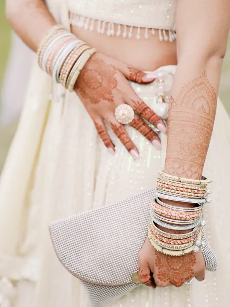 Fotografia de detalhe Fine Art das mãos de uma noiva indiana com desenhos Mehndi profundos, a segurar uma bolsa elegante rodeada de joias tradicionais.