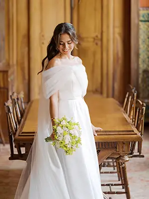 Smiling bride in Lisbon wearing an off-the-shoulder wedding dress and holding a white and green floral bouquet, in a classic interior with wood paneling.