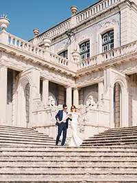 Newlyweds walking down the grand stone staircase of a historical palace in Lisbon under a clear blue sky.