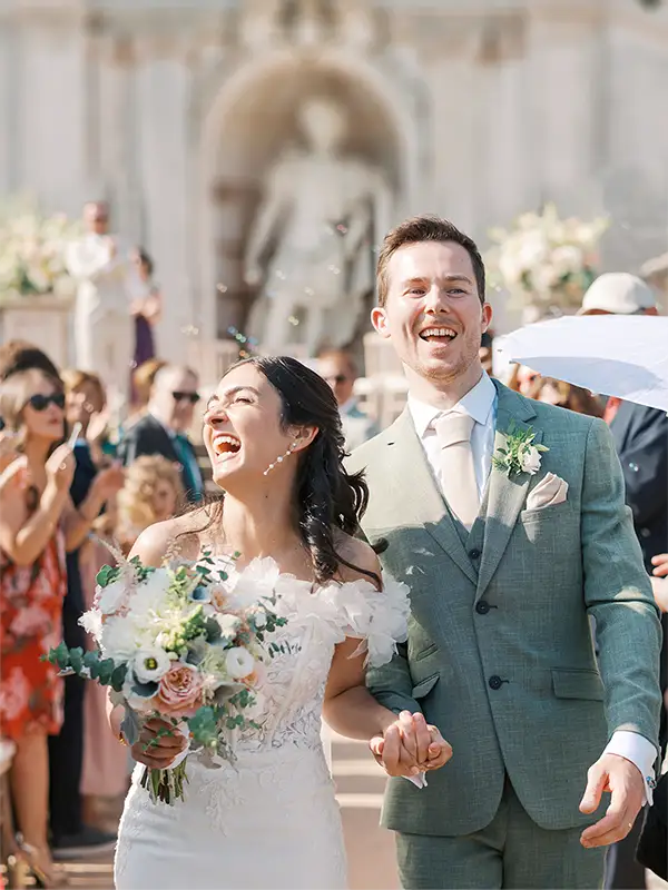 Newlyweds Nicole and Richard joyfully celebrate their wedding processional in Lisbon, among guests and petals, featuring a detailed floral bouquet.